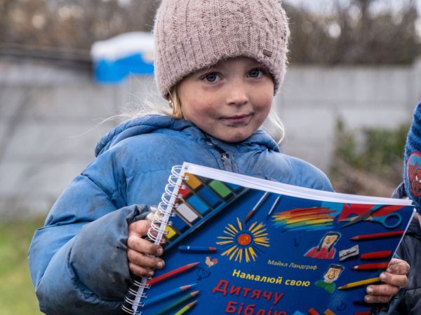 Ein ukrainisches Mädchen in Daunenjacke und Mütze hält eine Kinderbibel zum Selbstgestalten in der Hand.